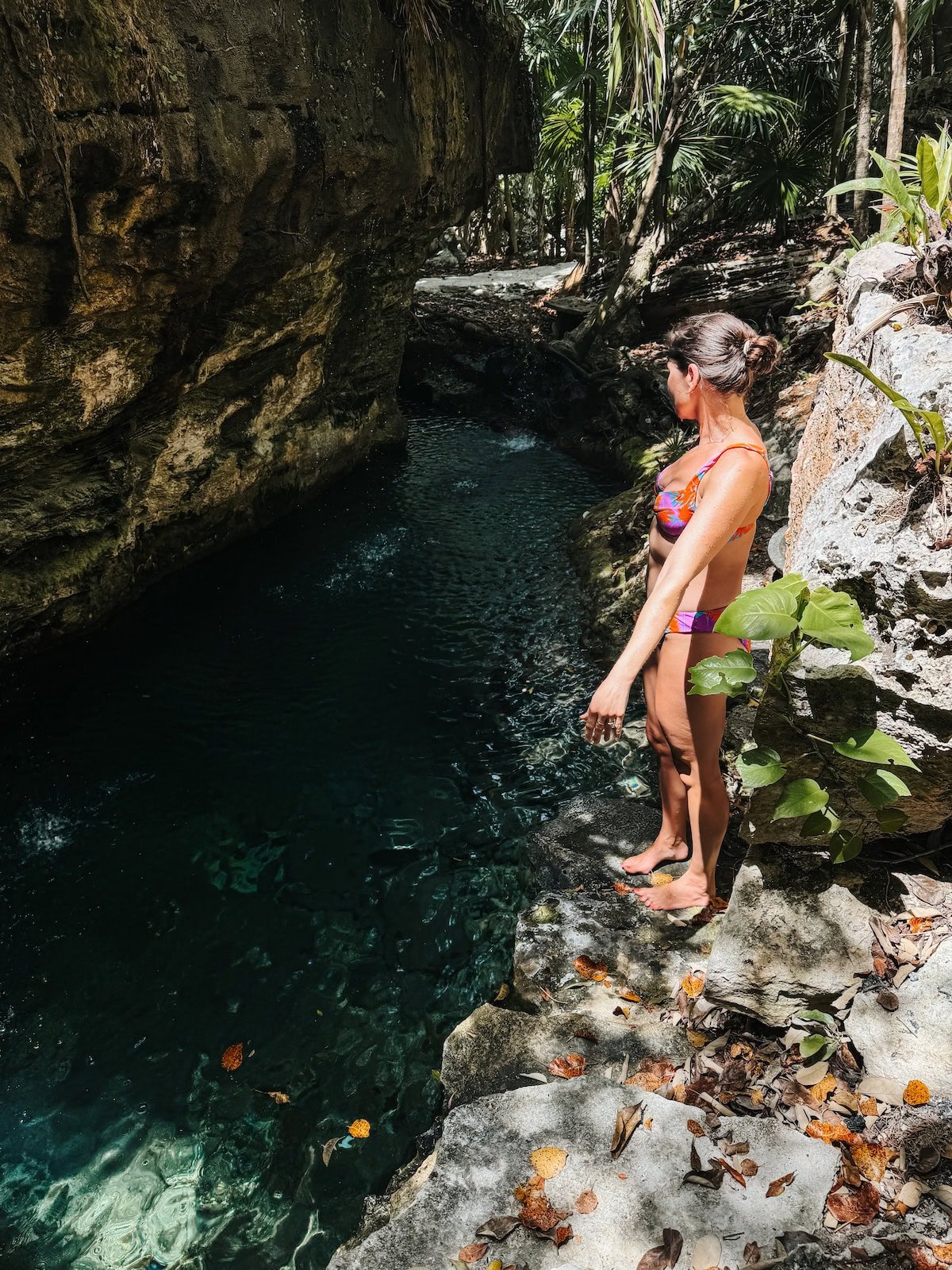 A woman in a swimsuit stands on rocky ground near a clear, forested cenote.