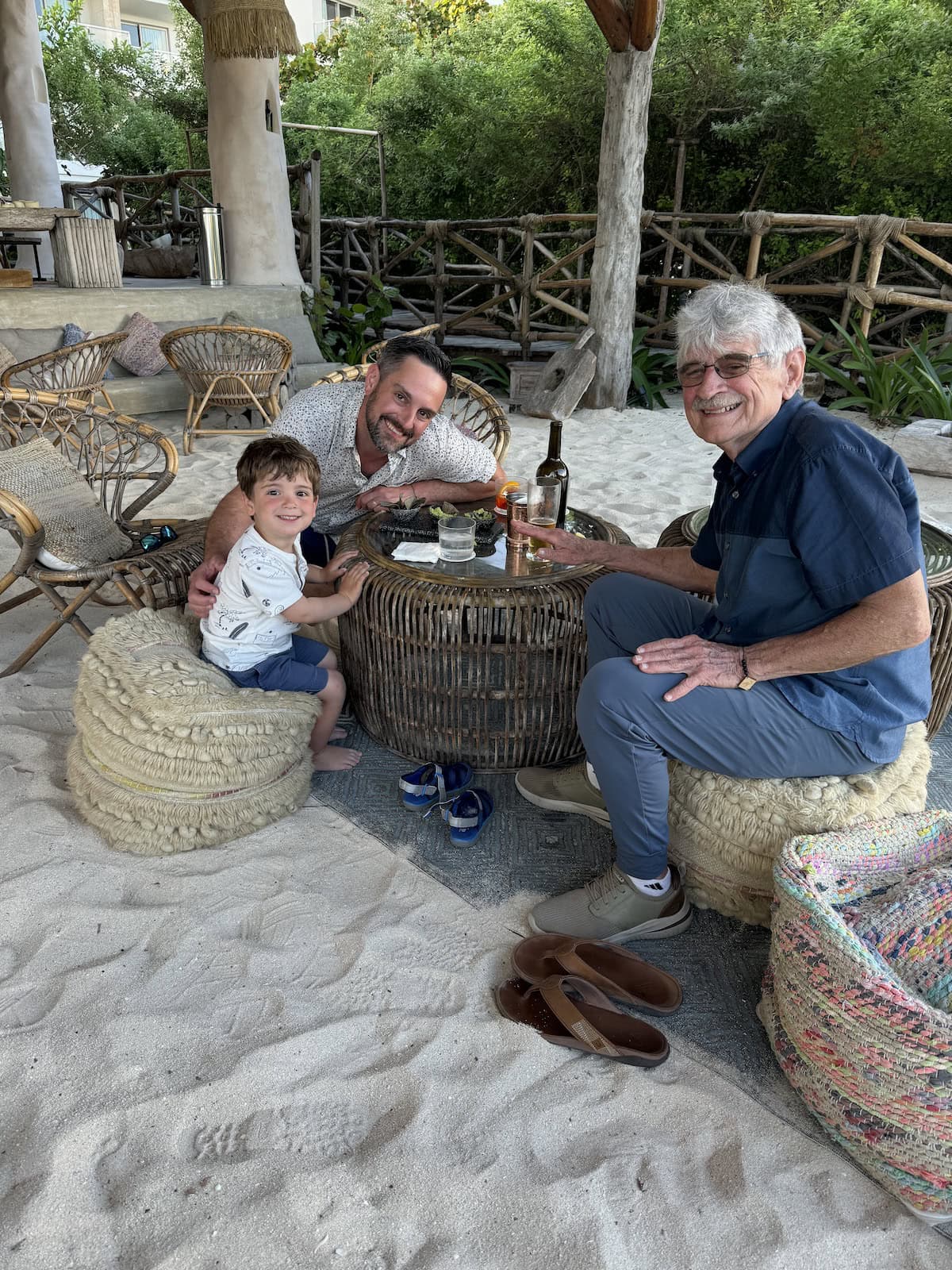 A man, child, and older man smiling and sitting at a low table on the sand.