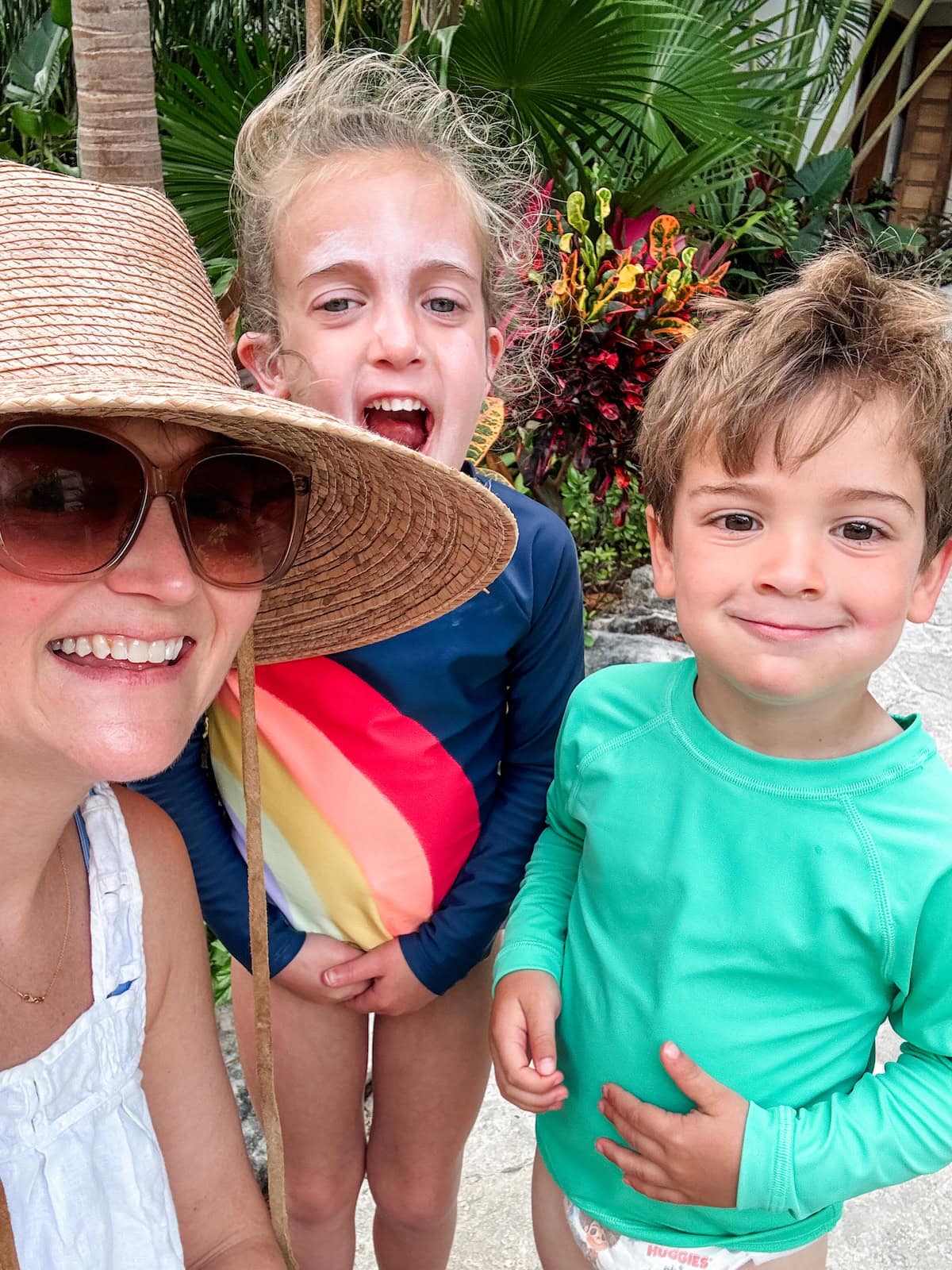 A woman and two children smiling outdoors, dressed for swimming.