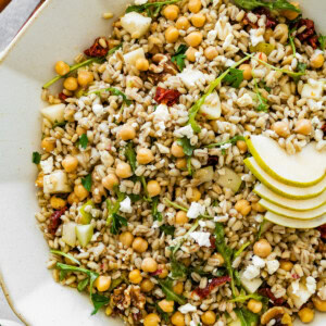 Large serving bowl filled with barley salad mixed with chickpeas, arugula, pear slices, sun-dried tomatoes, walnuts, and crumbled feta with wooden serving spoons on the side.