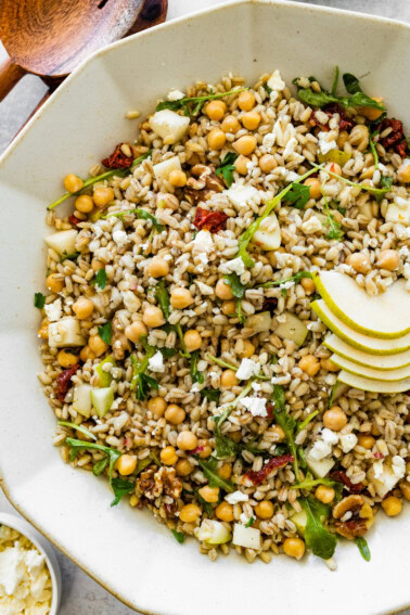 Large serving bowl filled with barley salad mixed with chickpeas, arugula, pear slices, sun-dried tomatoes, walnuts, and crumbled feta with wooden serving spoons on the side.