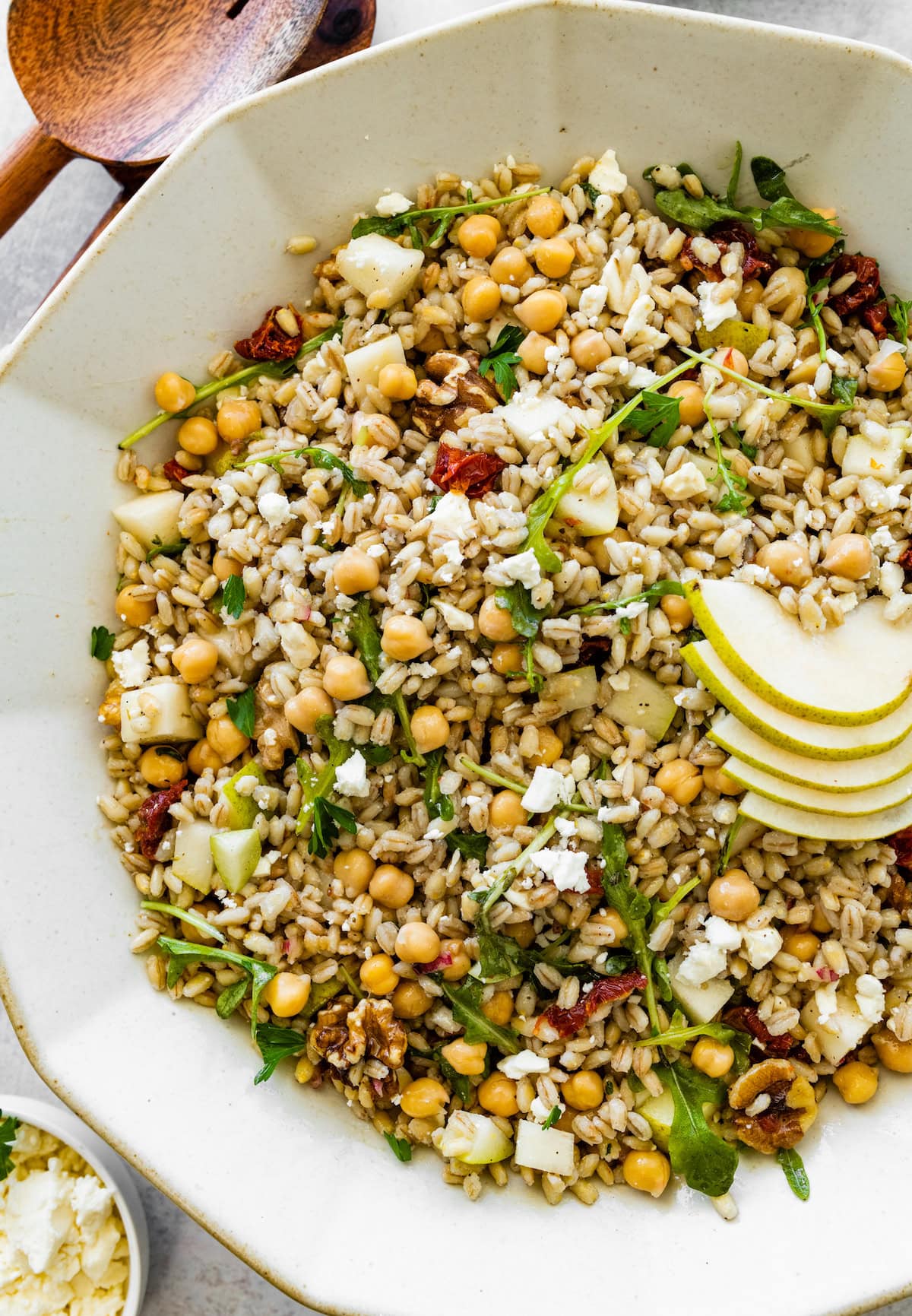 Large serving bowl filled with barley salad mixed with chickpeas, arugula, pear slices, sun-dried tomatoes, walnuts, and crumbled feta with wooden serving spoons on the side.