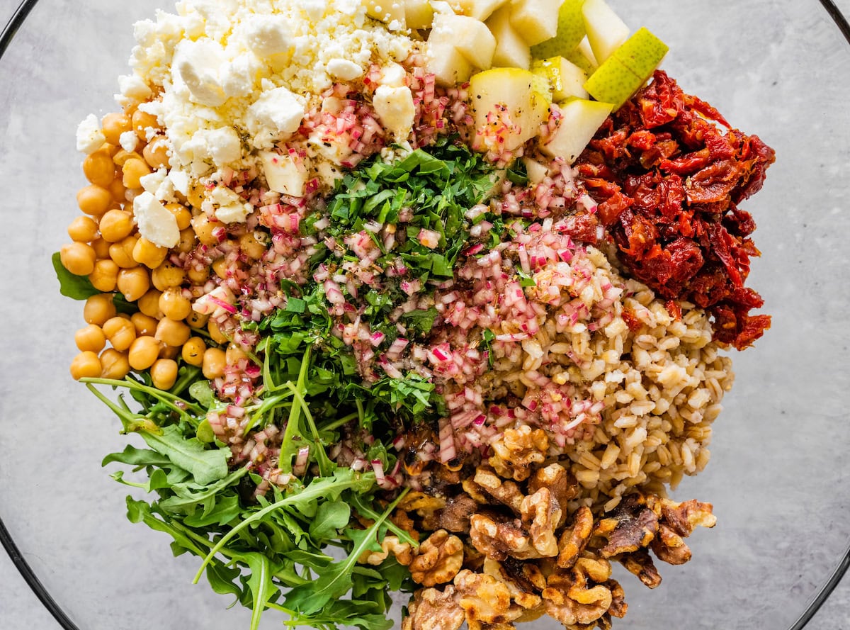 A glass mixing bowl with barley, chickpeas, pears, walnuts, sun-dried tomatoes, feta, and chopped herbs.