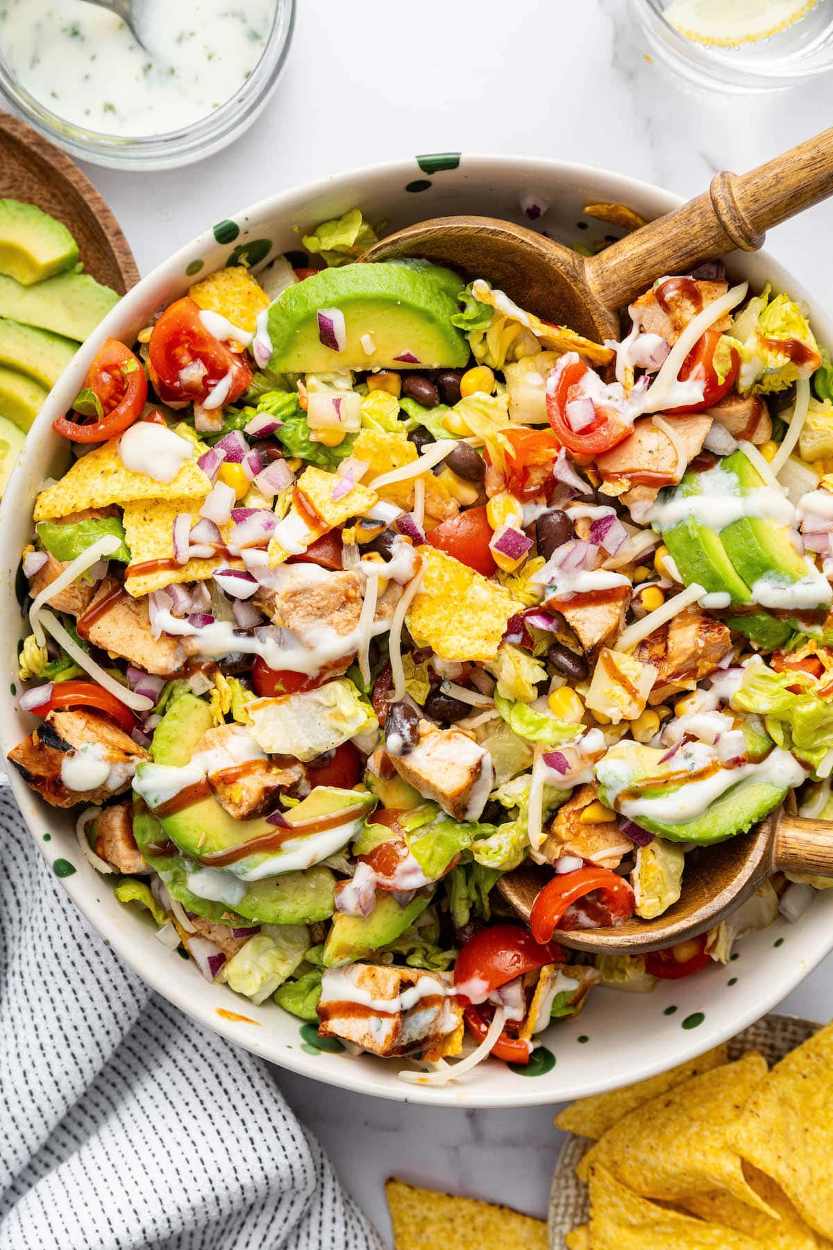 A BBQ chicken salad with avocado slices, BBQ chicken, corn, tortilla chips, tomatoes, lettuce, black beans all topped with a drizzle of BBQ sauce and ranch dressing. There are wooden salad spoons in the salad.