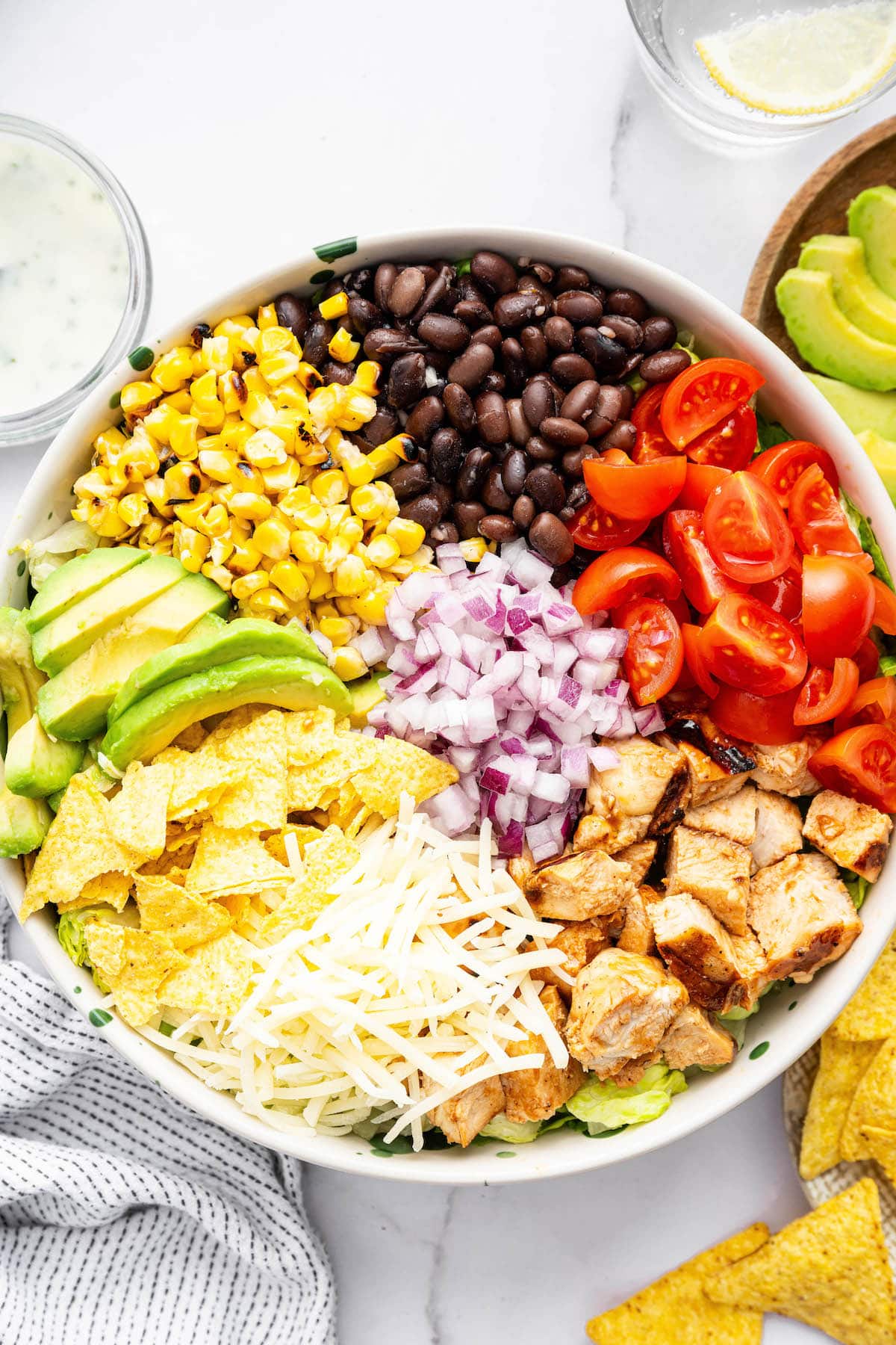 Ingredients for the BBQ chicken salad sectioned off on top of lettuce: red onions, black beans, cherry tomatoes, BBQ chicken, shredded cheese, avocado, corn and tortilla chips.