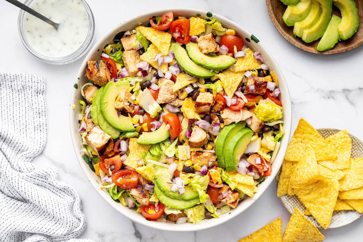 A BBQ chicken salad with avocado slices, BBQ chicken, corn, tortilla chips, tomatoes, lettuce and black beans.