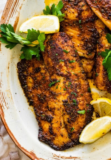 Blackened tilapia filets on a plate topped with fresh parsley and served with lemon wedges.