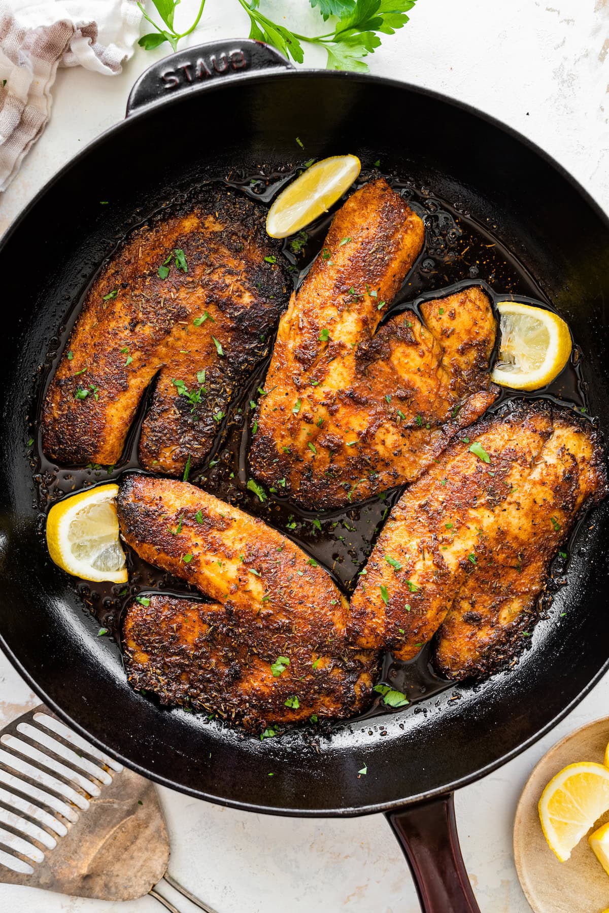 Four blackened tilapia filets cooking in a cast iron skillet topped with fresh parsley. There are lemon wedges in the skillet.