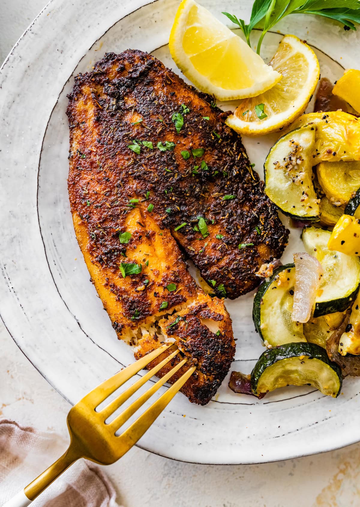 A blackened tilapia filet on a plate topped with fresh parsley and served with lemon wedges and roasted squash.