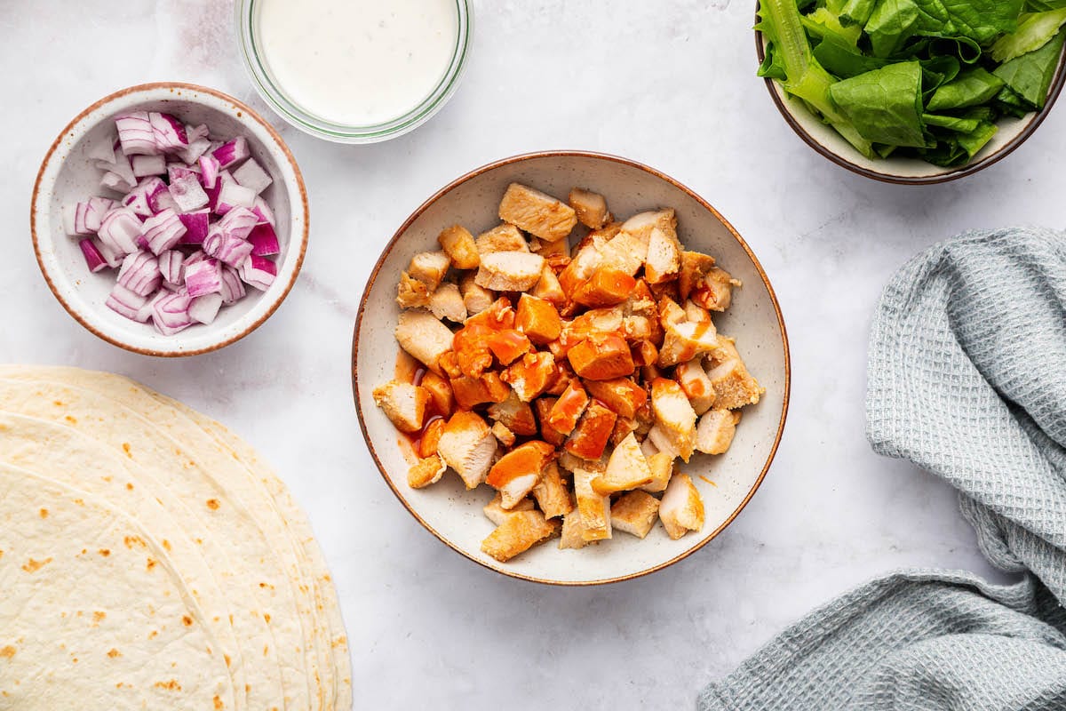 Bowl of chopped chicken tenders tossed in buffalo sauce with toppings in small bowls nearby.