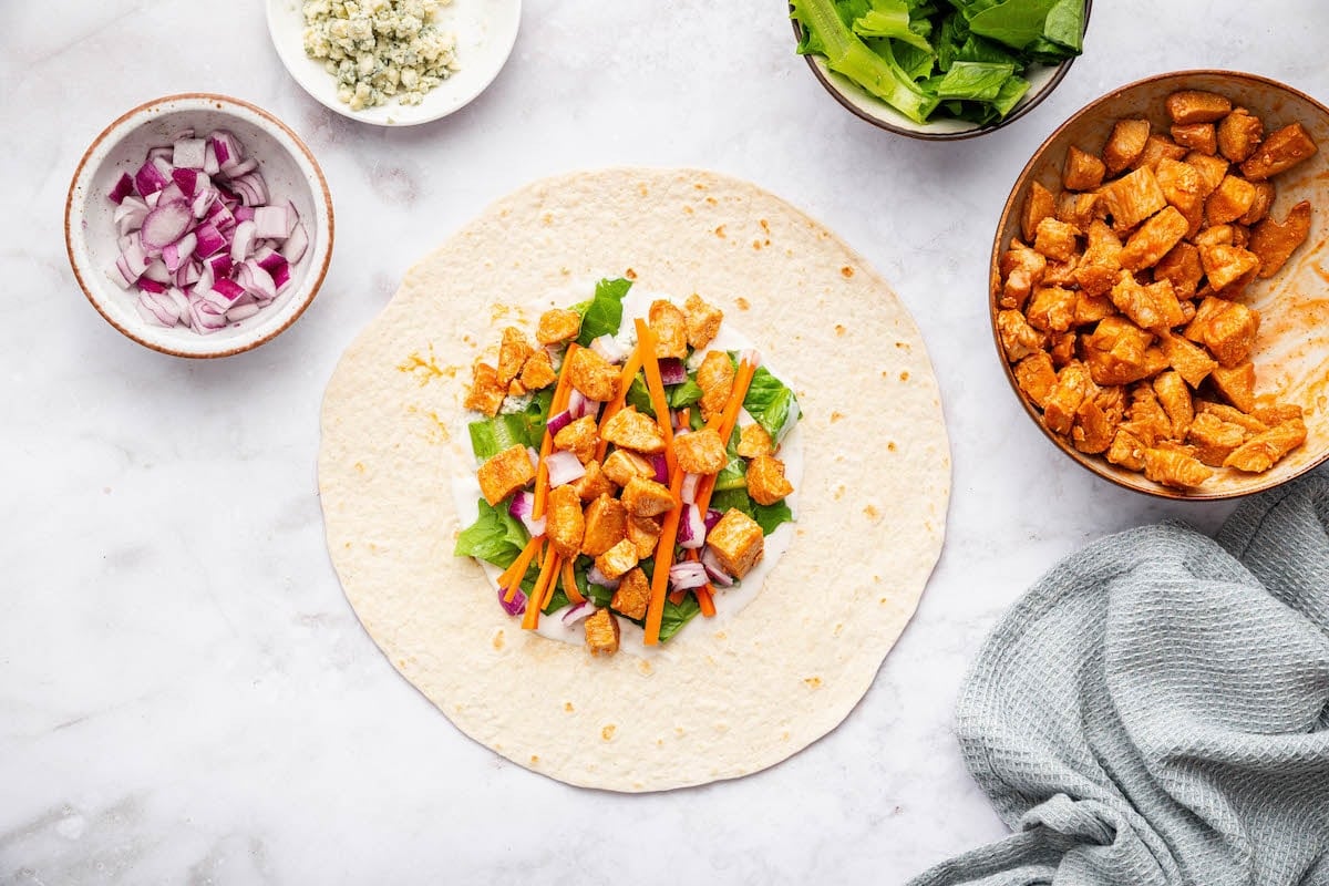 Flour tortilla topped with buffalo chicken, romaine lettuce, carrot strips, and red onion, ready to wrap.