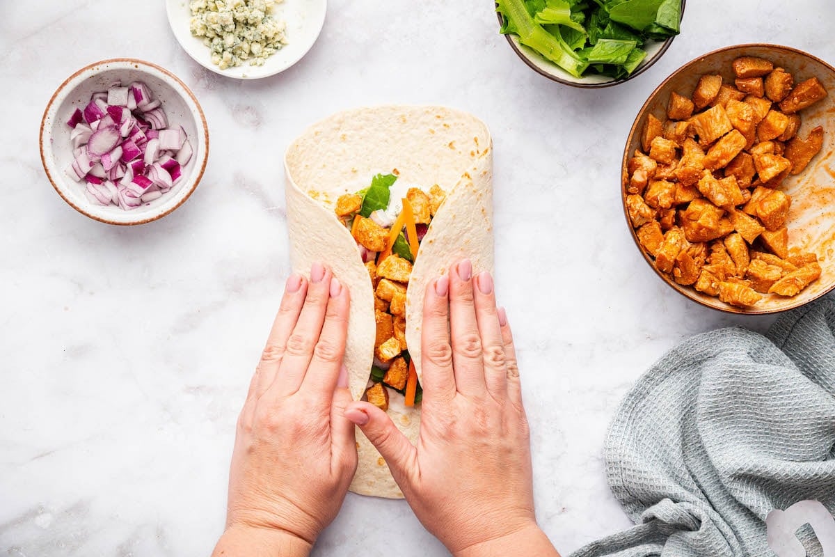Hands folding a tortilla filled with buffalo chicken and toppings to make a wrap.