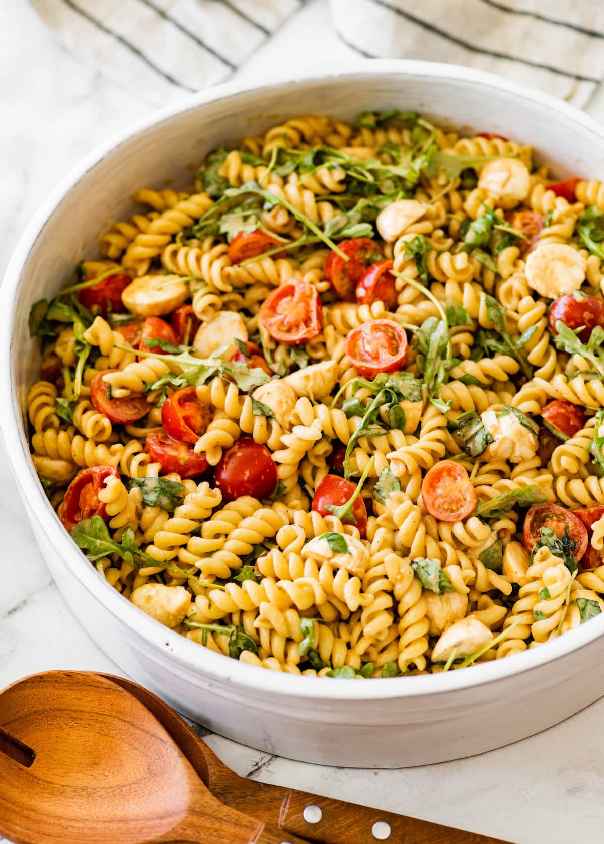 A serving bowl filled with caprese pasta salad. There are serving spoons next to the bowl.