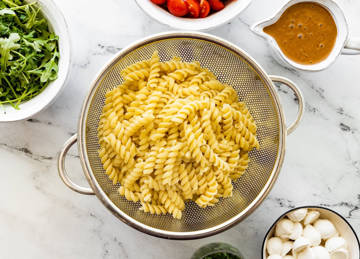 Cooked pasta in a strainer. There is a bowl of dressing next to the strainer.