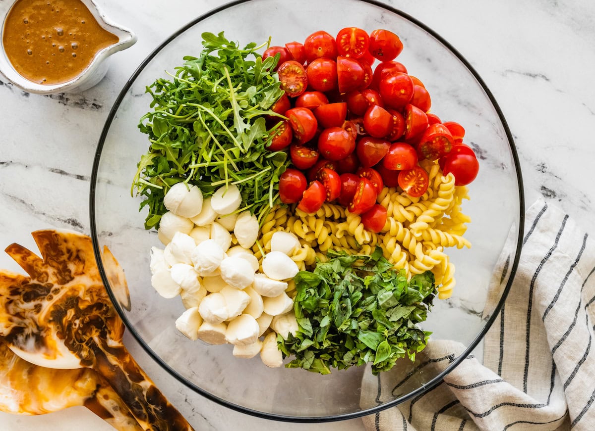 Ingredients for the caprese pasta salad in a glass mixing bowl: cherry tomatoes, arugula, mozzarella, basil and pasta.