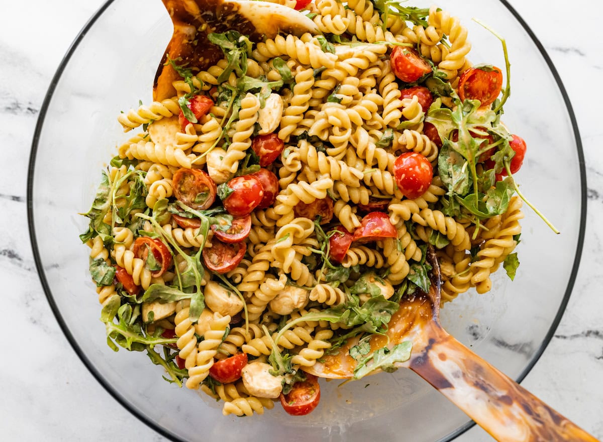 Caprese pasta salad in a glass mixing bowl being mixed with serving spoons.