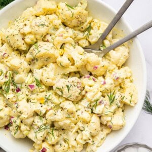 Cauliflower potato salad in a large serving bowl with silver serving utensils. The salad is topped with fresh dill.