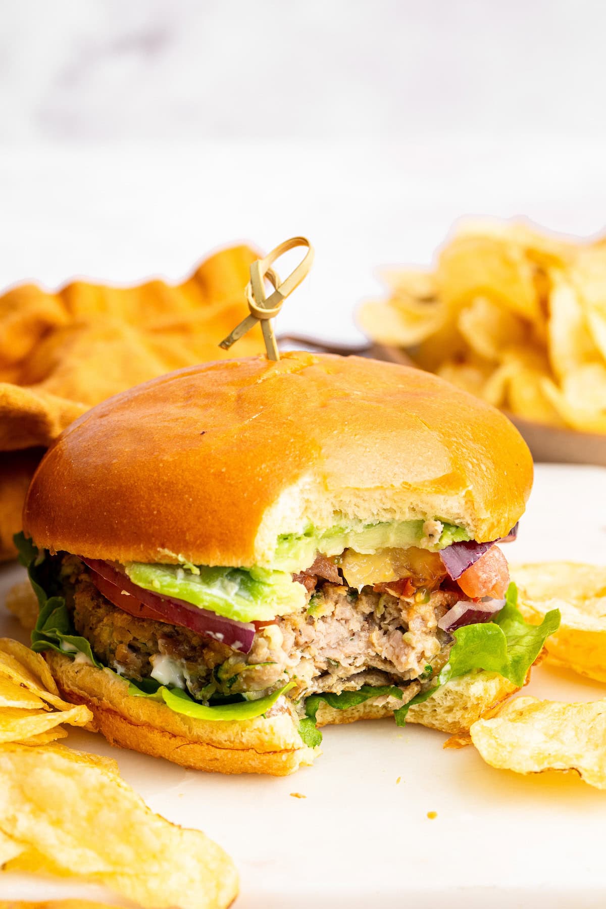 A chicken burger served on a brioche bun topped with lettuce, tomato, red onion and avocado. The burger is served with a side of potato chips.