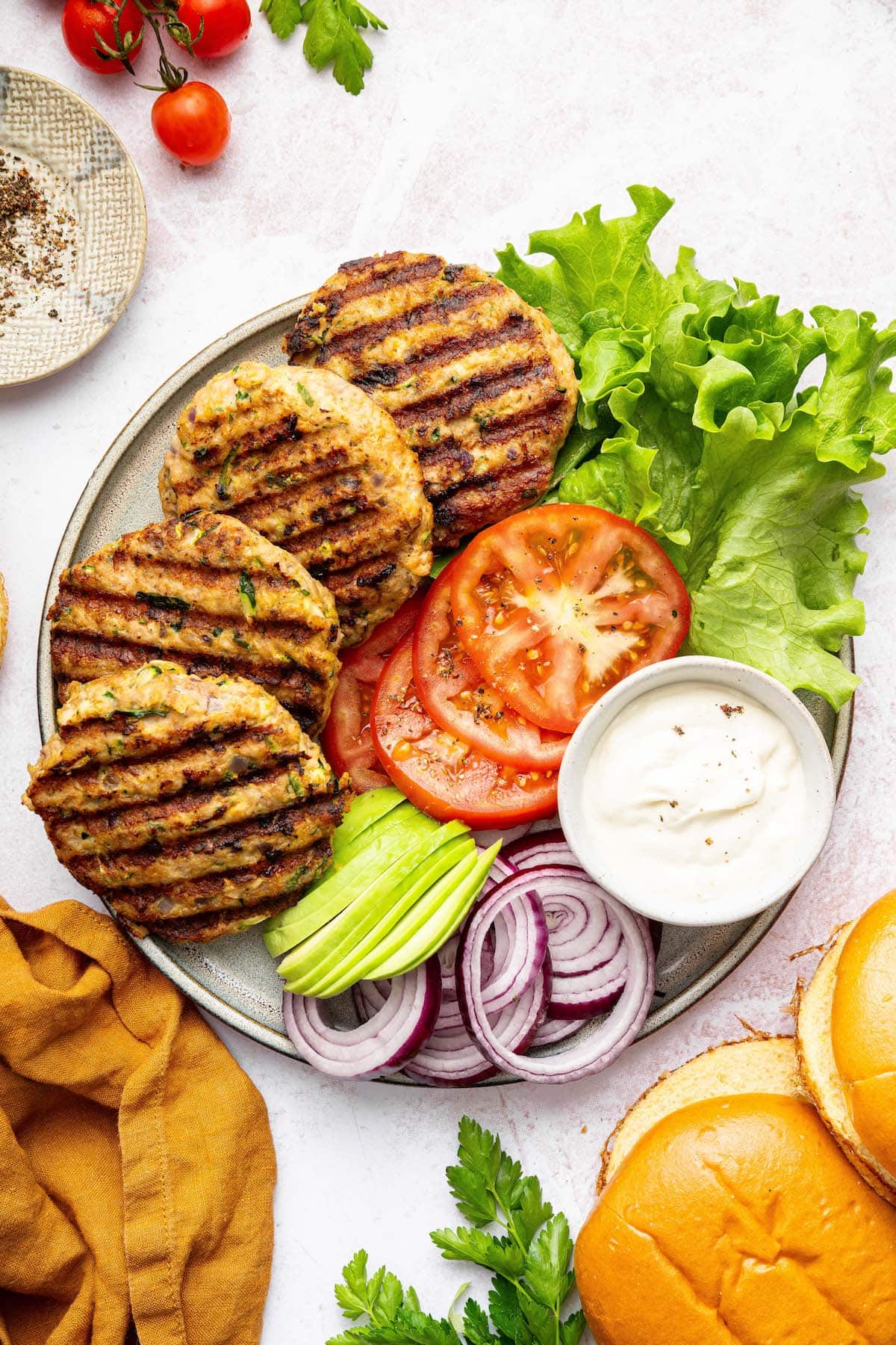 Four chicken burger patties on a plate with sliced tomatoes, red onion, avocado, lettuce and a garlic aioli.