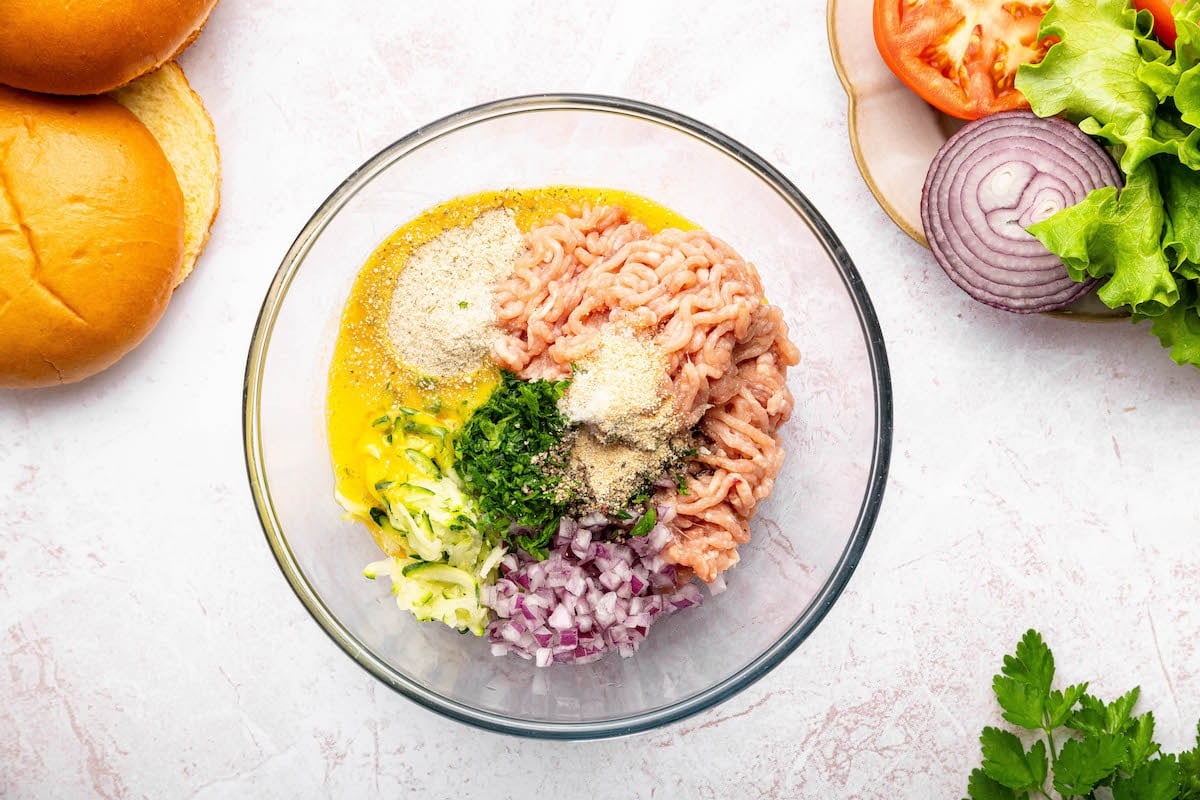 The chicken burger ingredients in a glass bowl: ground chicken, parsley, red onion, shredded zucchini, breadcrumbs, egg, and seasonings.