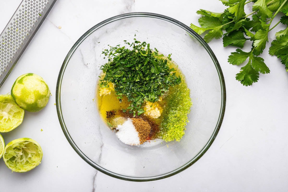 The cilantro lime marinade ingredients in a glass bowl: lime juice, lime zest, cilantro, salt, pepper, garlic, olive oil, maple syrup, cumin and paprika.