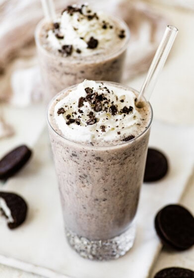 Two cookies and cream protein shakes in glasses, topped with whipped cream and crushed chocolate sandwich cookies, with clear straws inserted.
