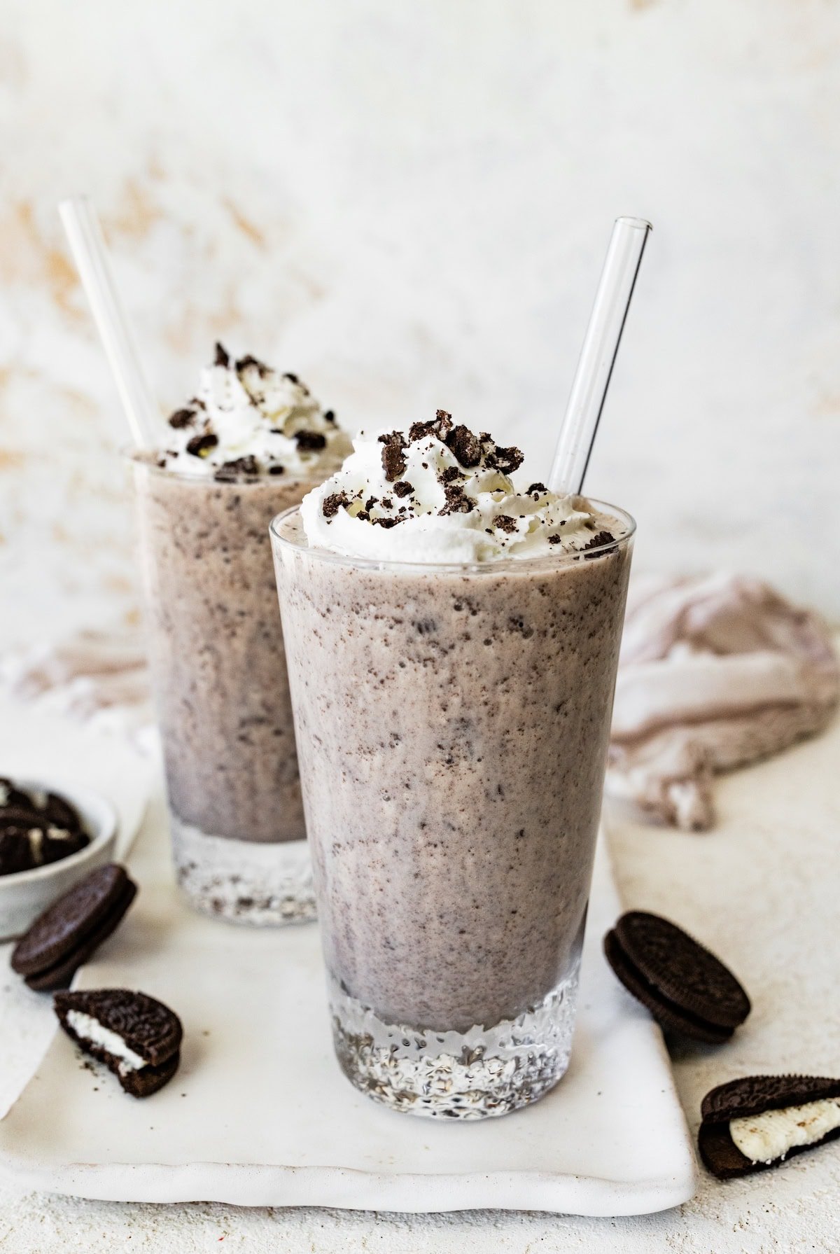 Two glasses of cookies and cream protein shake topped with whipped cream and cookie crumbles, with whole sandwich cookies scattered around.