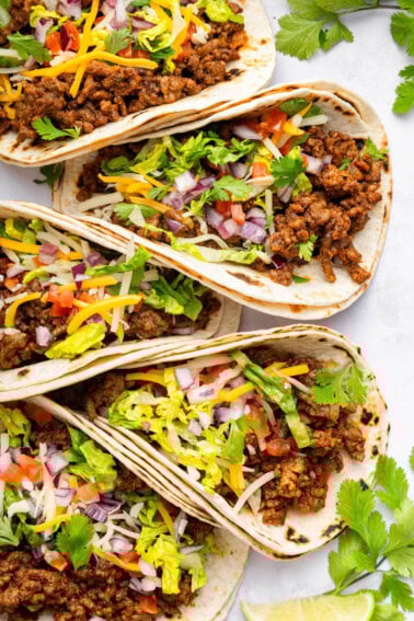 Five ground beef tacos in flour tortillas topped with lettuce, onion, tomato, cilantro and cheese.