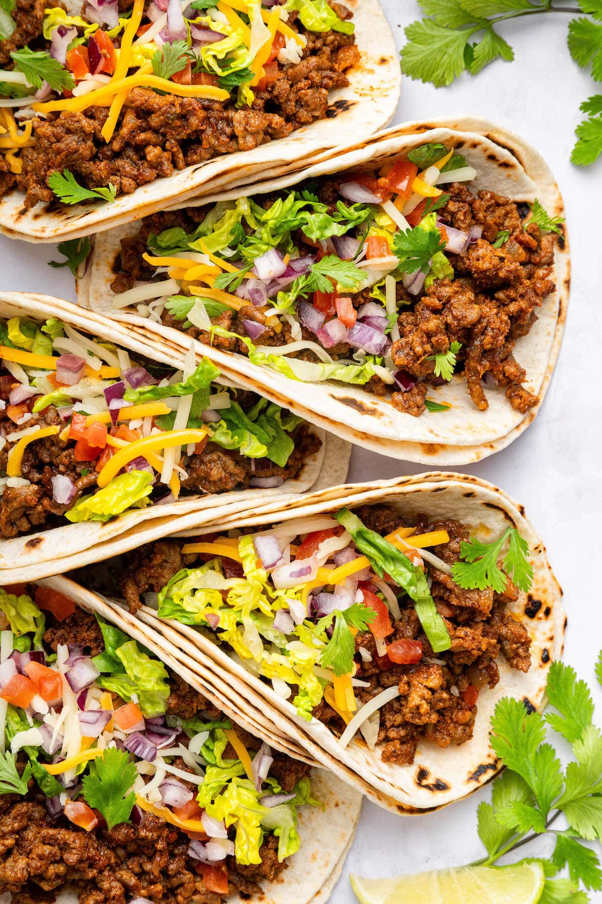 Five ground beef tacos in flour tortillas topped with lettuce, onion, tomato, cilantro and cheese.