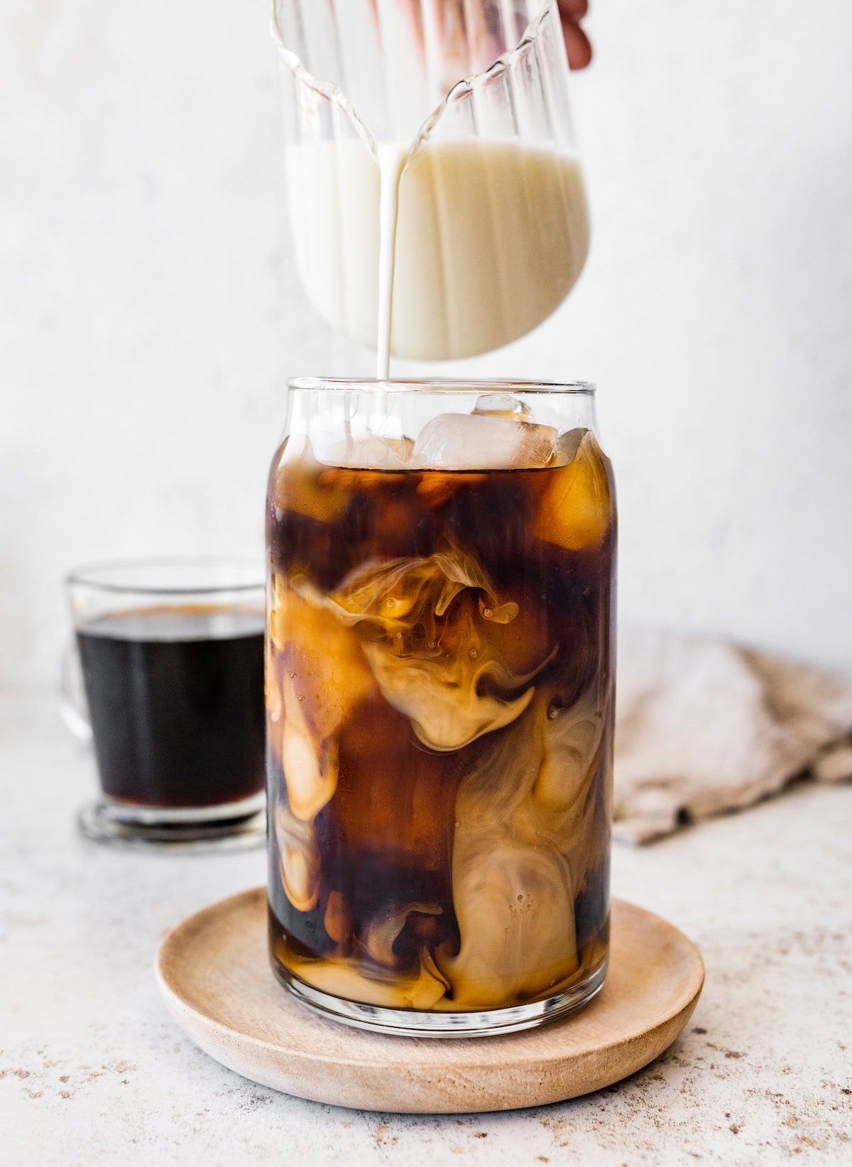 Milk being poured from a clear glass pitcher into a glass of iced coffee, creating swirling patterns as it mixes.