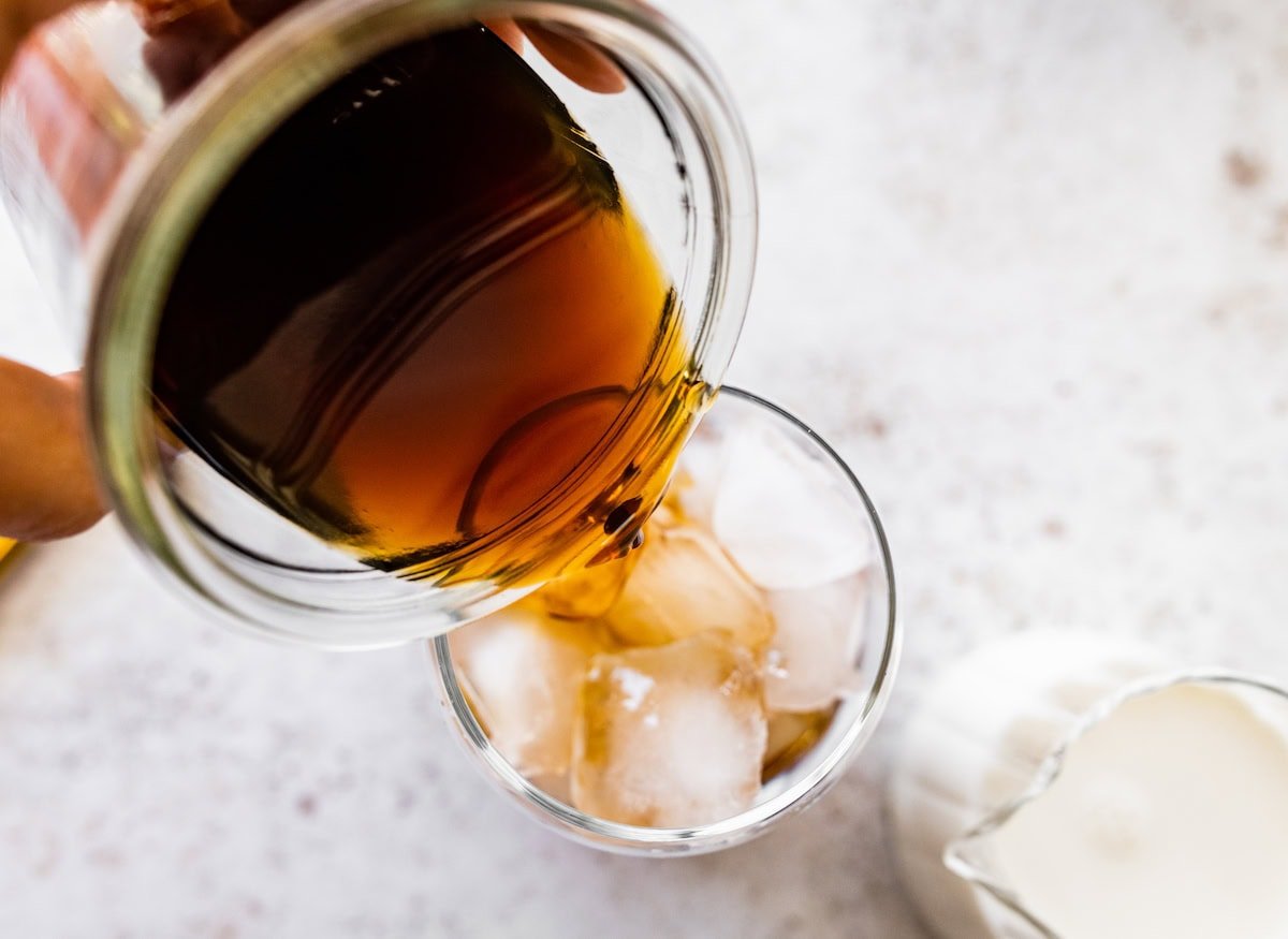 Black coffee being poured into a glass filled with ice, with milk in a pitcher nearby.