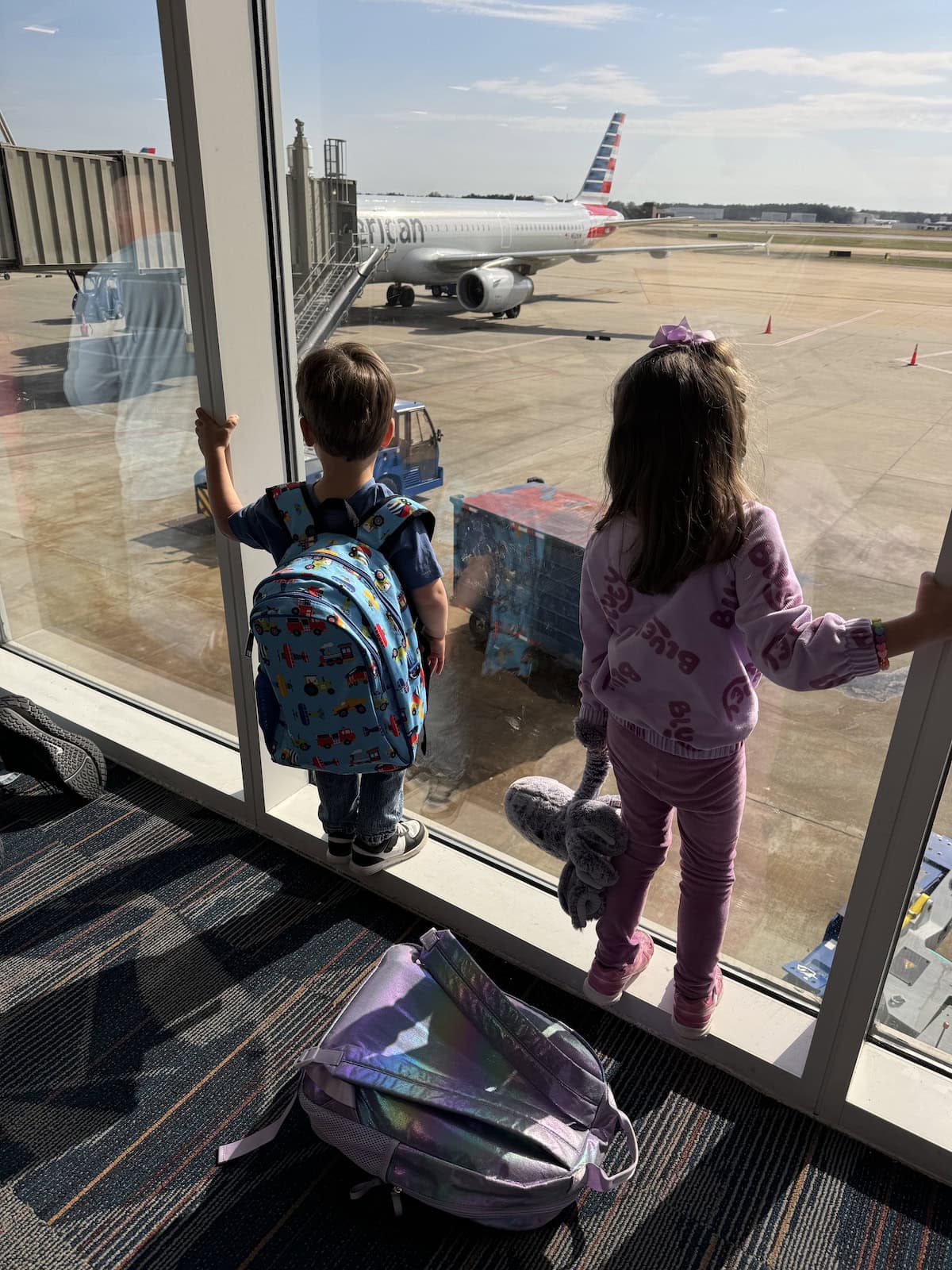 Two young children look out an airport window at an American Airlines plane.
