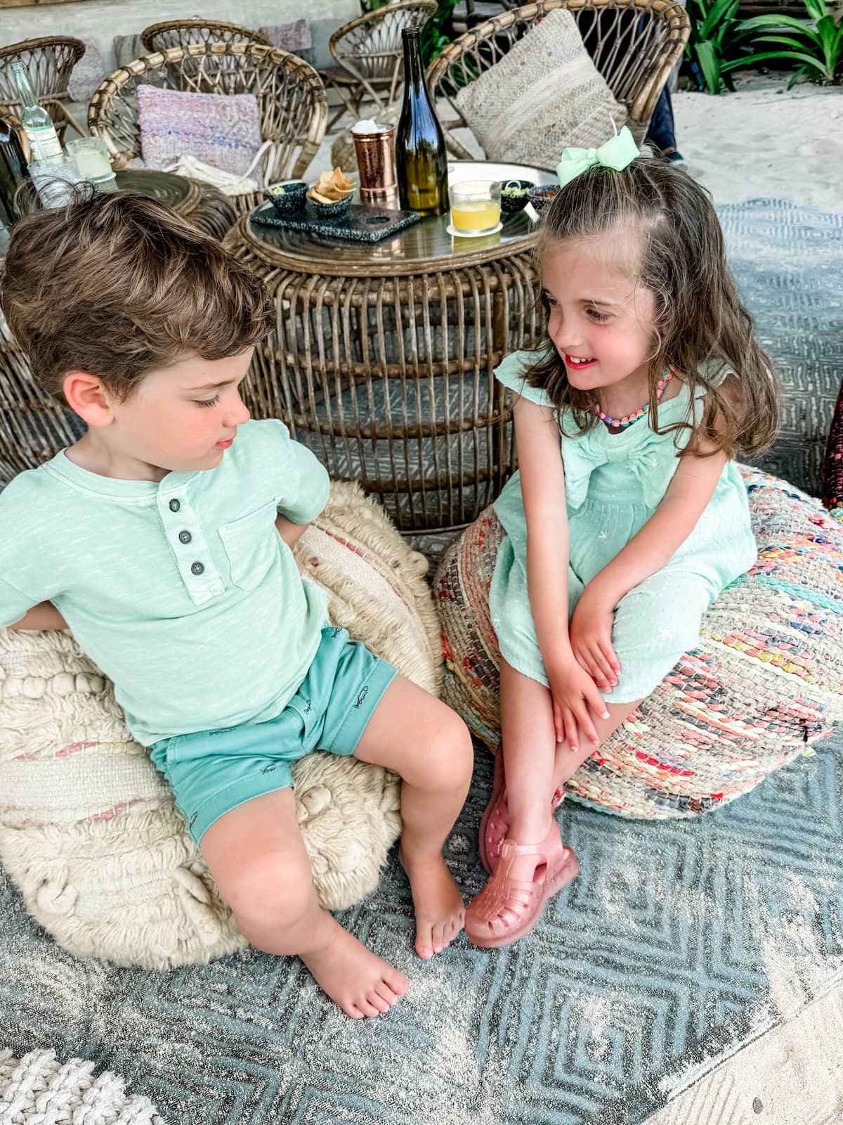 Two children sitting on small bags near a table on a sandy beach.