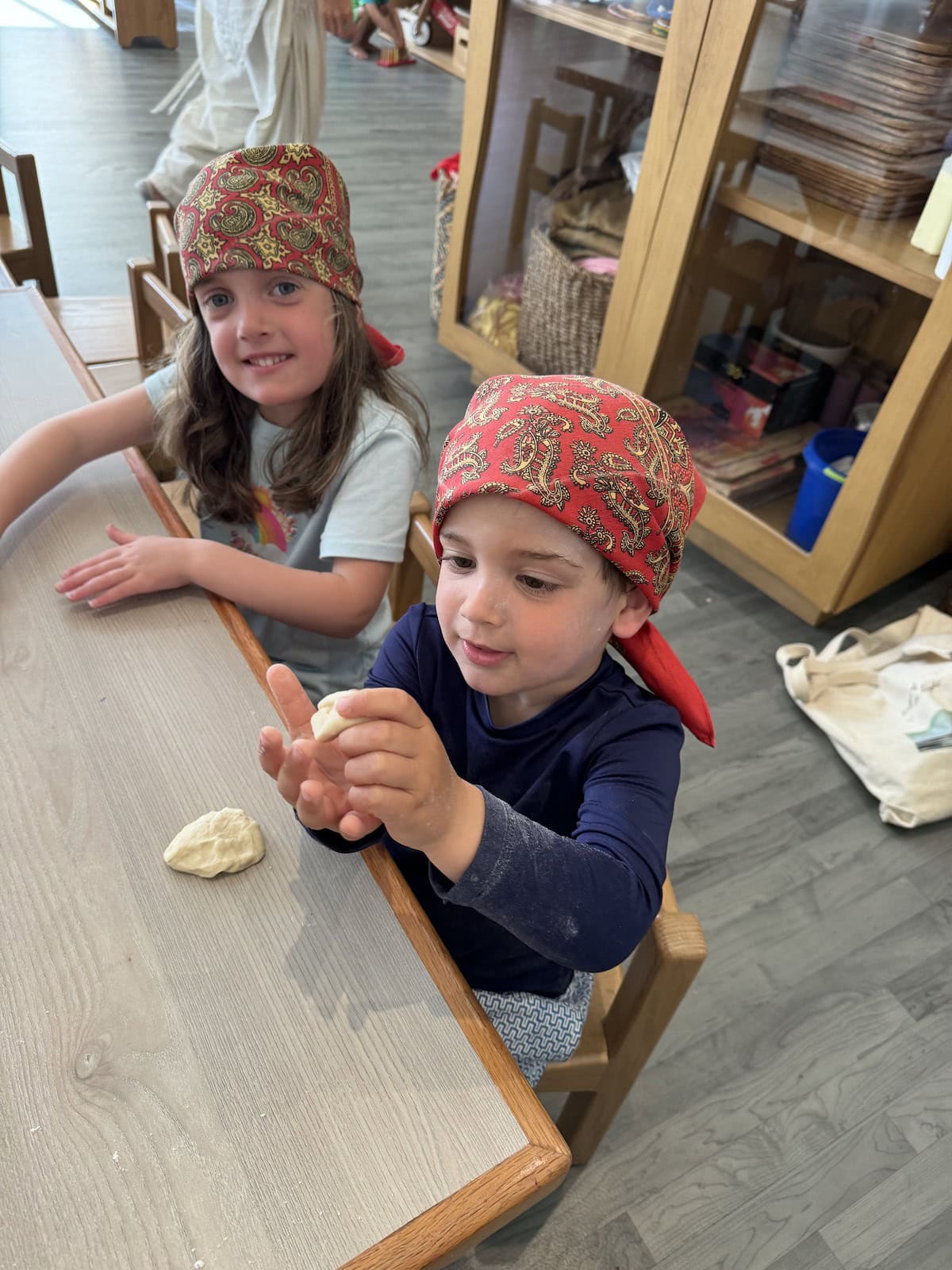 Two young children wearing matching red bandanas sit at a table shaping dough with their hands.
