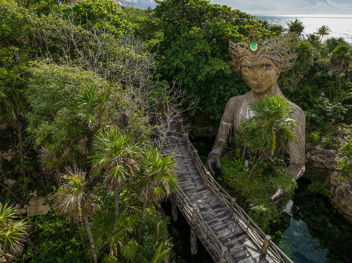 Large nature-inspired sculpture of a woman surrounded by jungle with a wooden bridge leading to it.
