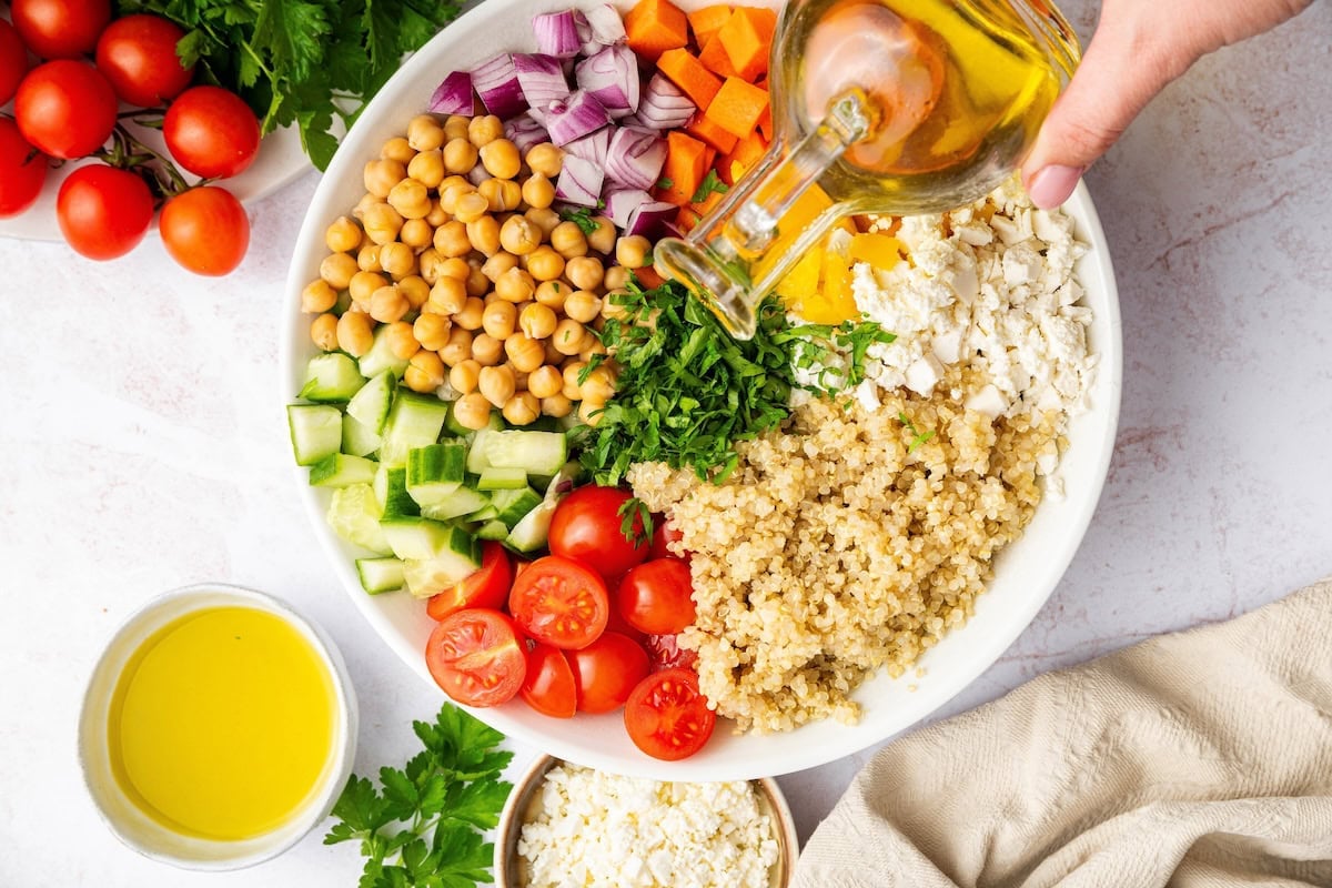 Pouring olive oil into a bowl of salad ingredients including quinoa, chickpeas, and vegetables.