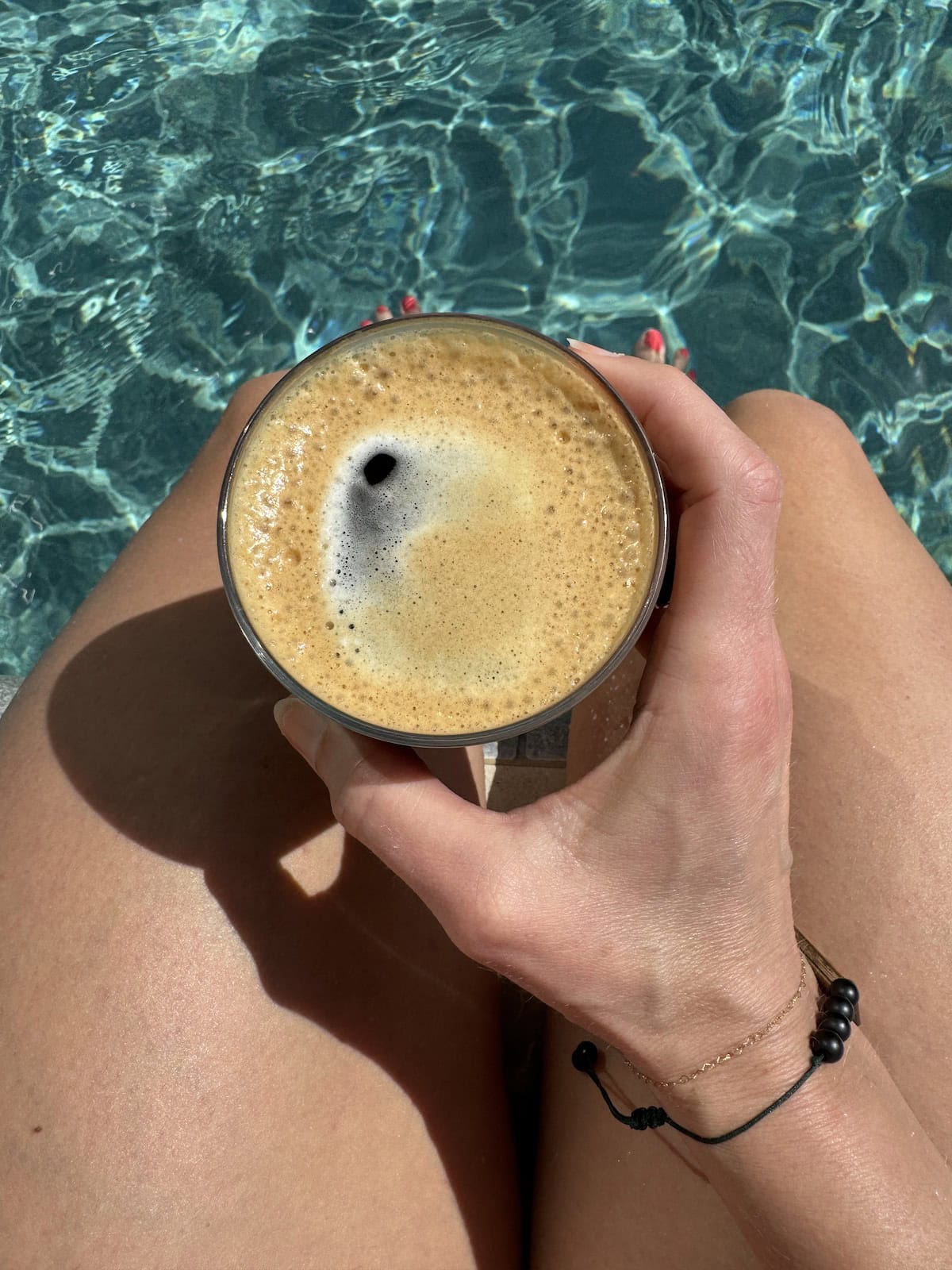A hand holding a glass of frothy coffee over sunlit legs and a sparkling pool.