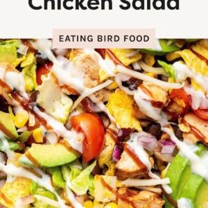 Colorful BBQ chicken salad topped with avocado, cherry tomatoes, corn, black beans, tortilla chips, shredded cheese, and drizzles of ranch and BBQ sauce.