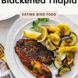 Blackened tilapia fillets cooking in a cast iron skillet with vegetables and lemon wedges on the side