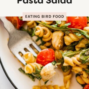 Close-up of a fork holding Caprese pasta salad with rotini, cherry tomato, mozzarella, and arugula on a white plate.