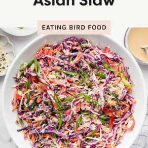 Colorful bowl of crunchy Asian slaw topped with sliced scallions and sesame seeds, shown with creamy dressing on the side.