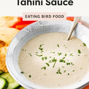 Bowl of tahini sauce topped with parsley, surrounded by fresh veggies and pita chips.
