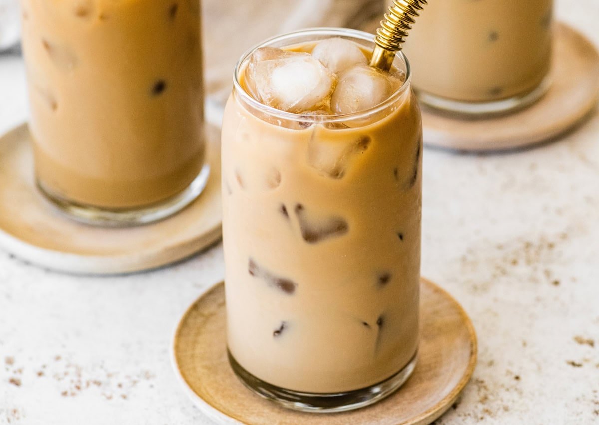 A glass of iced coffee with milk fully mixed in, served on a small wooden plate with a metal straw.