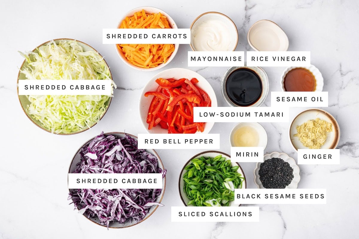 Crunchy Asian Slaw ingredients: Shredded cabbage, shredded carrots, mayonnaise, rice vinegar, sesame oil, low-sodium tamari, red bell pepper, mirin, ginger, black sesame seeds, sliced scallions.