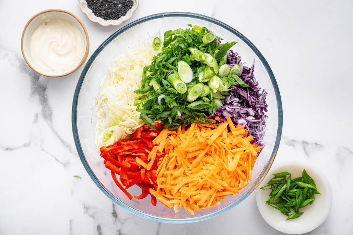 A glass mixing bowl filled with colorful prepped veggies for Asian slaw, including shredded green and purple cabbage, grated carrots, red bell pepper, and sliced green onions. Small bowls of black sesame seeds, dressing, and extra scallions sit nearby.