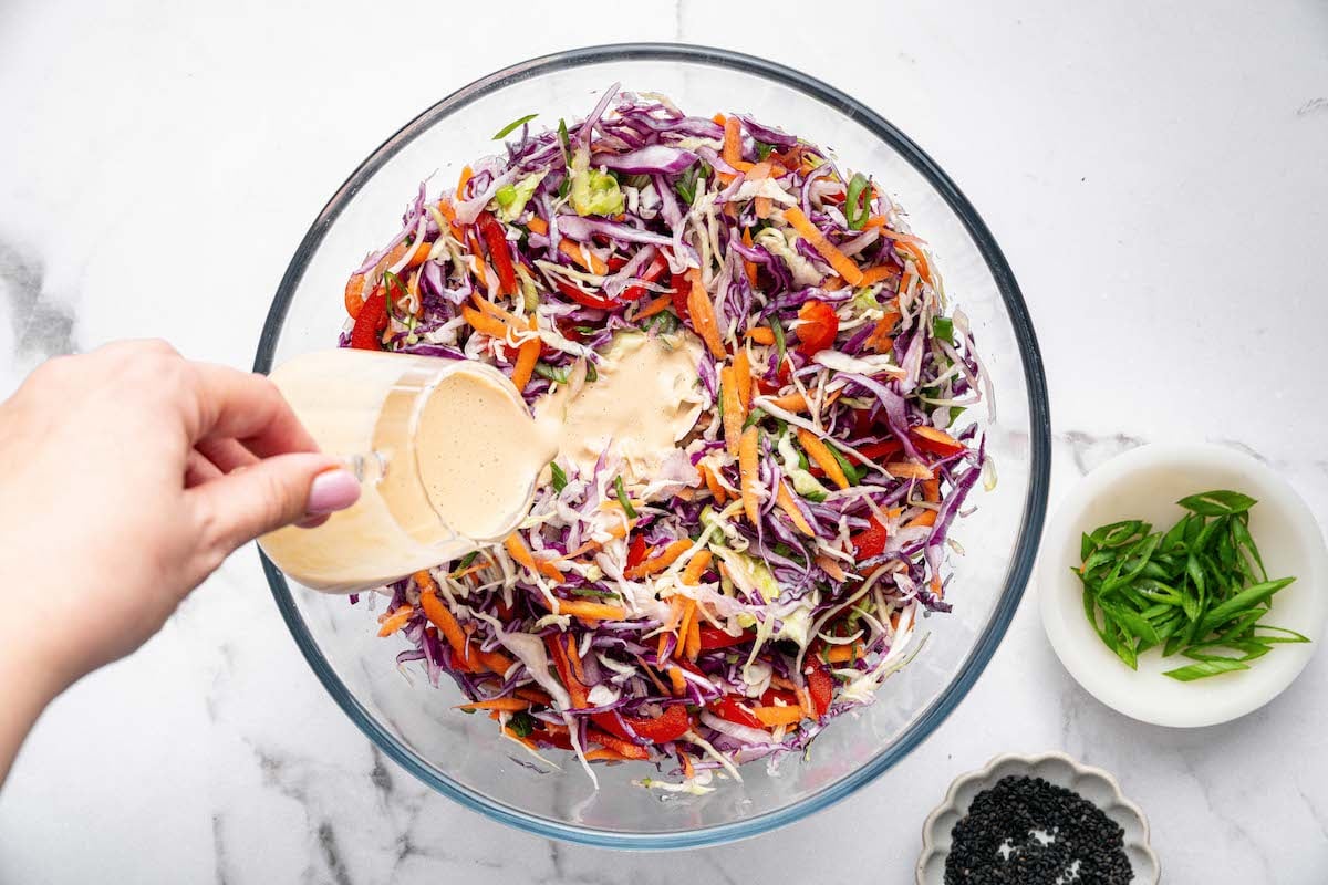 Hand pouring creamy sesame dressing over a large bowl of shredded cabbage slaw with carrots, red pepper, and scallions. A small bowl of sesame seeds and green onions are on the side.