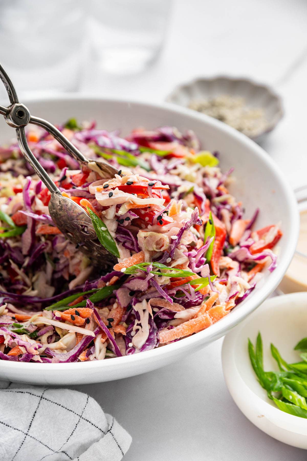 Tongs lifting a serving of colorful Asian slaw from a bowl, revealing the creamy texture and mix of crunchy vegetables and sesame toppings.