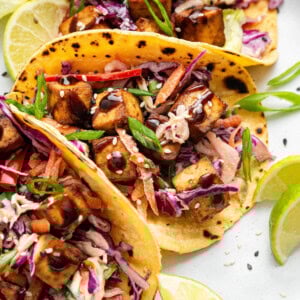 Three tofu tacos in yellow corn tortillas, loaded with slaw, tofu cubes, scallions, and sesame seeds, with lime slices in the background.