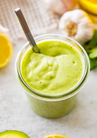 Glass jar filled with avocado dressing, garnished with parsley, lemon, and garlic in the background.