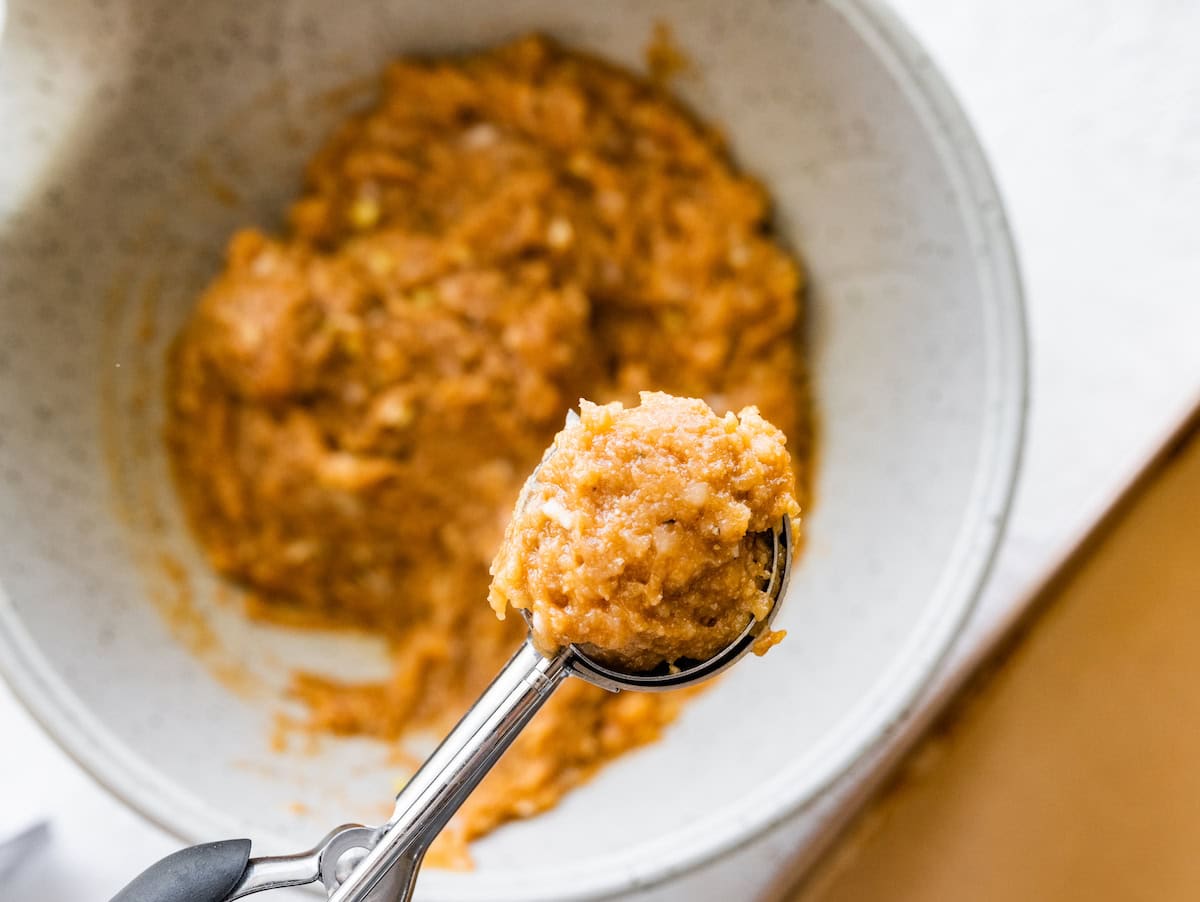 Scooping raw chicken meatball mixture from a bowl with a cookie scoop.